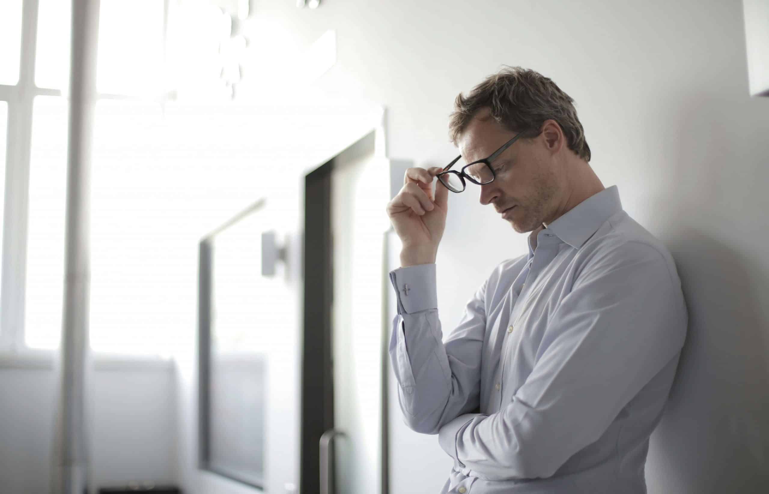A man in a smart, white shirt, leaning on an office wall, holding his glasses and looking at the floor, as if suffering from eye strain.