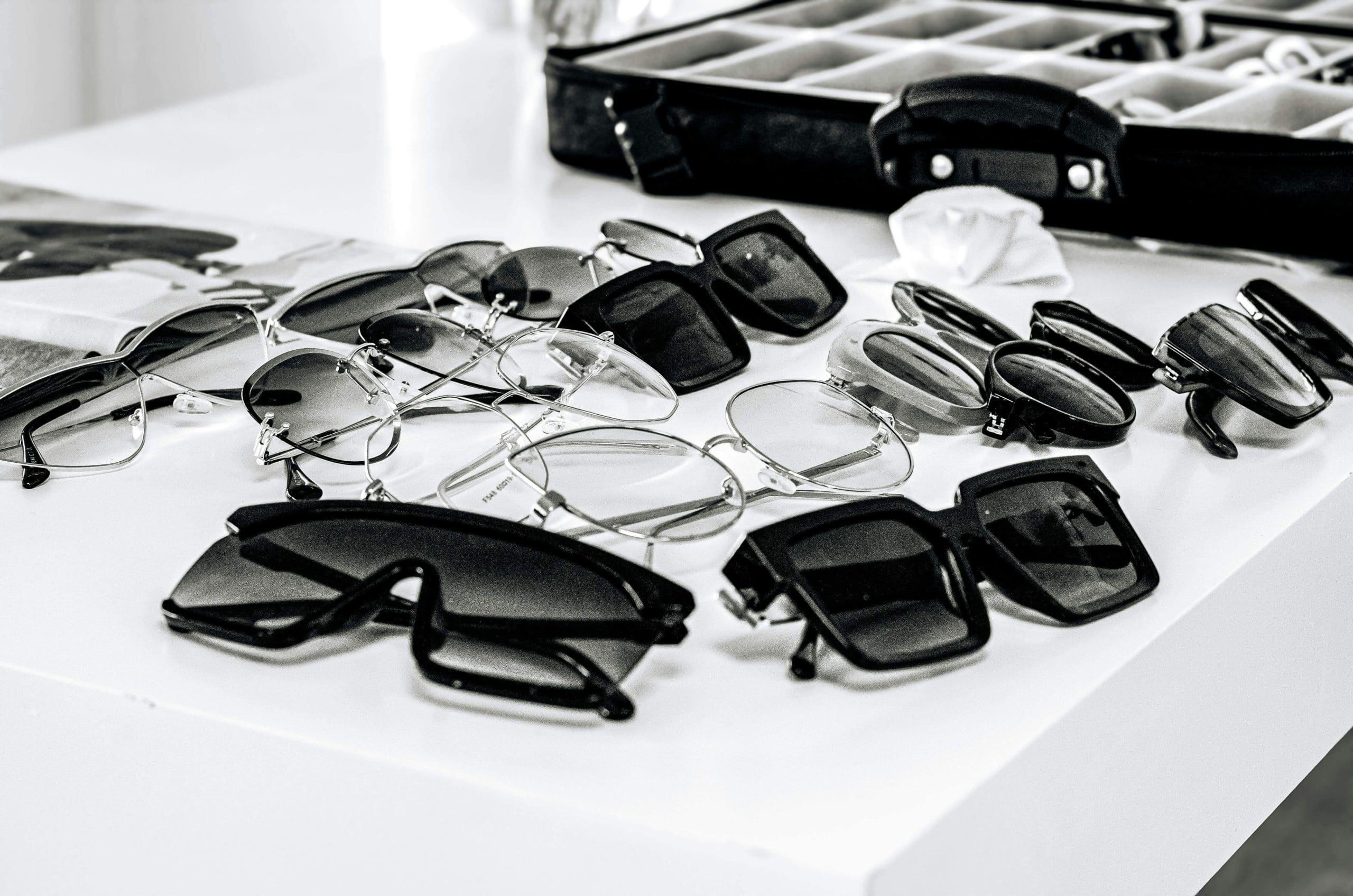 Black-and-white image of various glasses and sunglasses scattered on a desk, representing the different types of lenses for glasses.